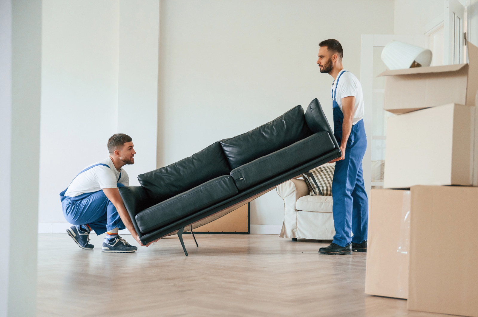 Two movers lift a black sofa off an oak herringbone parquet floor in a Stanmore home, clearing the space for Dust-Free Floor Sanding.