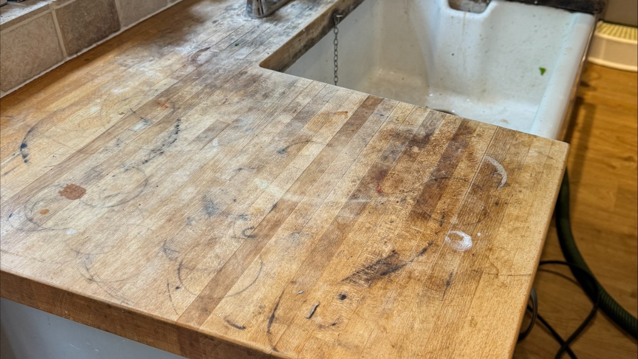 Heavily stained solid oak kitchen worktop beside a ceramic tile floor in a Stanmore home, shown before professional Worktop Refinishing.