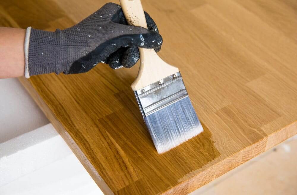 Gloved hand applying clear oil during Worktop Refinishing on a solid oak wooden kitchen worktop with matching oak hardwood floor in Stanmore.