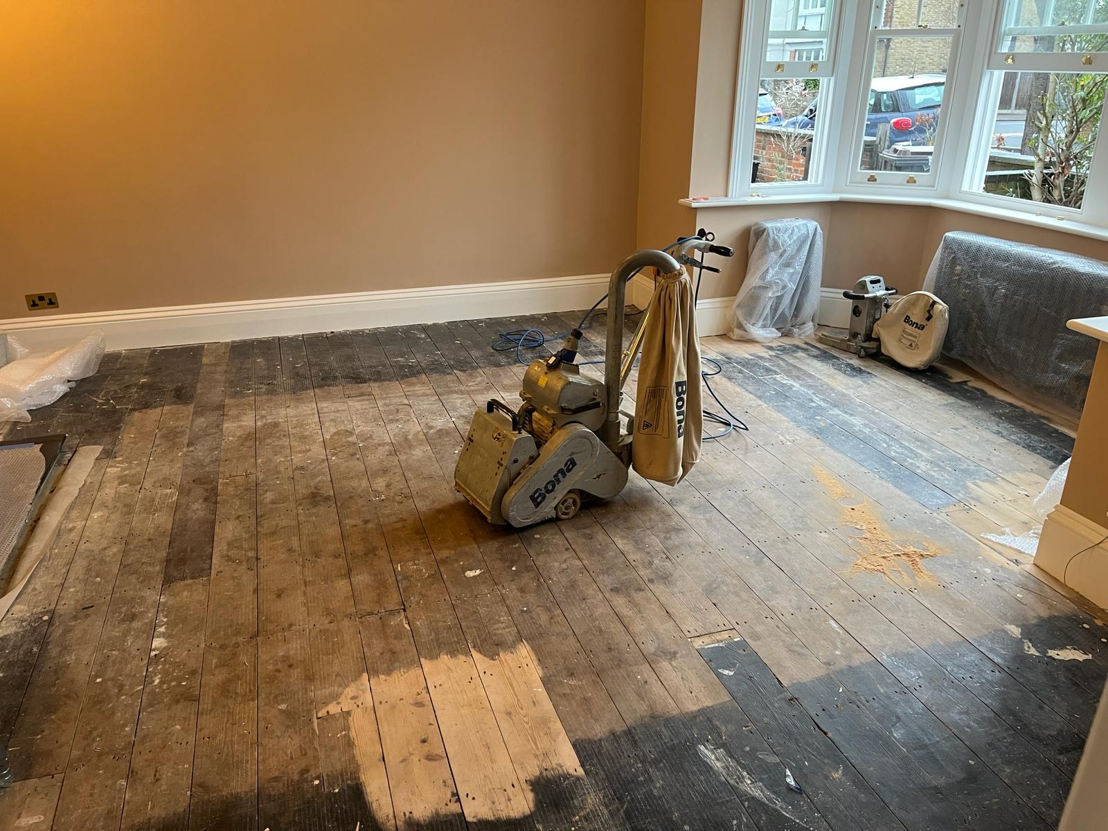 Wood Floor Sanding Stanmore – Original Pine Floorboards Restoration in Progress Wood Floor Sanding in Stanmore living room, showing Bona sanding machine stripping old finish from original pine floorboards for restoration.